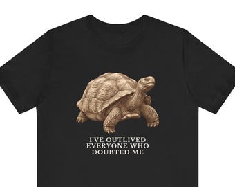 May include: A black short-sleeved t-shirt featuring a detailed illustration of a brown tortoise in the centre. Below the tortoise, the text reads: I'VE OUTLIVED EVERYONE WHO DOUBTED ME in white capital letters. The design is centred on the chest.