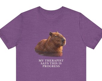 May include: A heather purple short-sleeve t-shirt featuring a photographic print of a brown capybara partially submerged in water. Below the animal, the text reads: MY THERAPIST SAYS THIS IS PROGRESS in white, capitalized, serif lettering.