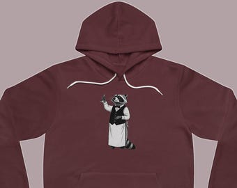 May include: A deep maroon pullover hoodie featuring a graphic print of a raccoon dressed as a waiter. The raccoon wears a black vest, a white apron, and a bow tie, holding a small glass. The hoodie includes a hood with white drawstrings.