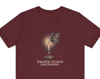 May include: A maroon short-sleeve t-shirt featuring a graphic of a moth hovering near a lit candle in a holder, set against a circular background. Below the graphic, the text reads "DRAWN TO BAD DECISIONS." in a white, serif font.