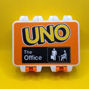 May include: A rectangular white and orange plastic case for a card game. The front features the word UNO in large yellow letters with black outlines. Below, a black rectangle contains the text The Office and a white icon of two figures.
