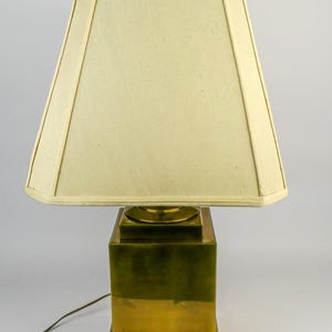 May include: A brass table lamp with a square base and a cream-colored lampshade. The lamp has a gold-colored finial on top. The lampshade is a trapezoid shape.