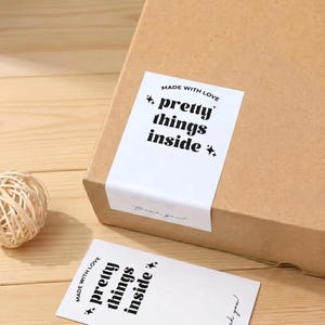 May include: A brown cardboard box with a white label that reads "MADE WITH LOVE, pretty things inside." A small, decorative ball of twine sits on a wooden surface next to the box. A second label is visible.