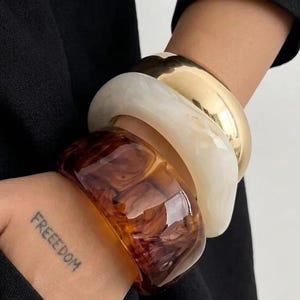 May include: A collection of four chunky bracelets in various colors and materials. The bracelets include a gold-tone metal, a creamy white marbled resin, and a translucent brown resin. The word "FREEDOM" is tattooed on the wrist.