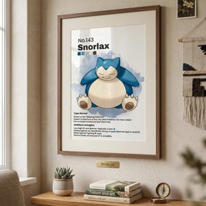 May include: Framed print featuring Snorlax, a blue and cream-colored Pok&eacute;mon, with the text "No.143 Snorlax." The print is in a wooden frame, displayed on a wooden shelf with books, a small plant, and a compass.
