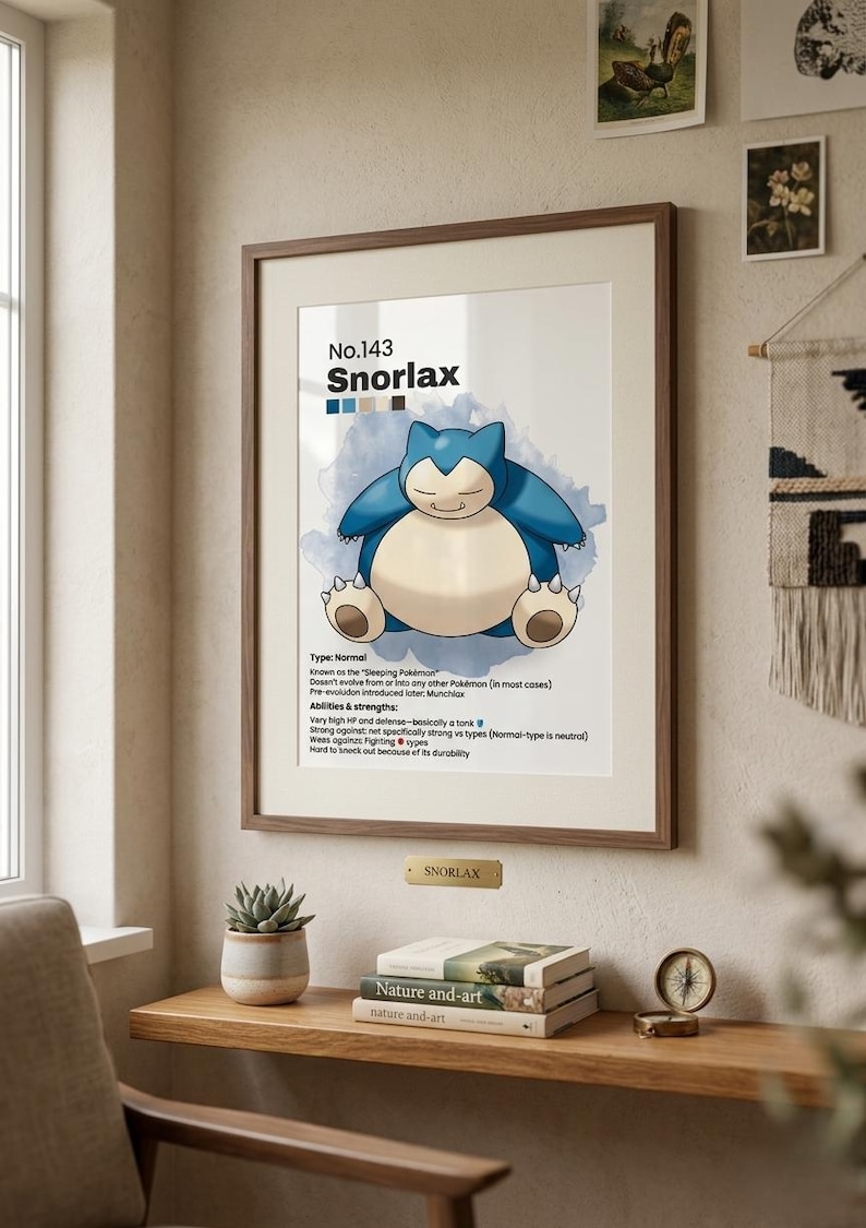 May include: Framed print featuring Snorlax, a blue and cream-colored Pok&eacute;mon, with the text "No.143 Snorlax." The print is in a wooden frame, displayed on a wooden shelf with books, a small plant, and a compass.