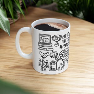May include: A white ceramic mug featuring black line art illustrations related to human resources. The design includes a laptop labeled HR PORTAL, a trophy, a handshake, a magnifying glass, a brain diagram, and the text Eat Hire Repeat.