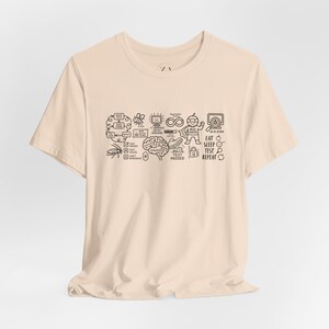 May include: A light beige short-sleeved t-shirt featuring a dark brown graphic print of software testing icons, including a brain, a bug, a superhero, and the text "EAT SLEEP TEST REPEAT" alongside various technical diagrams and flowcharts.
