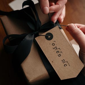 May include: A wrapped gift box with a black satin ribbon and a brown paper tag that says "open me". The gift box is wrapped in brown paper. The tag is attached with black twine.