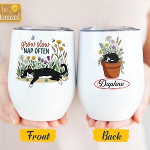 May include: Two white stainless steel tumblers with lids. The left tumbler features a black and white cat lying in a field of flowers with the text grow slow NAP OFTEN. The right tumbler shows a cat sleeping in a flower pot with the name Daphne.