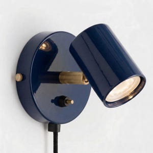 May include: A navy blue wall-mounted spotlight with a circular base and adjustable cylindrical head. The lamp features brass accents and a black power cord. The light is directed forward, illuminating the space.