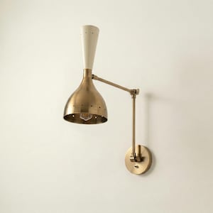 Könnte beinhalten: Eine Wandleuchte aus Messing mit weißem, konischem Oberteil und glockenförmigem Schirm. Die Lampe hat einen verstellbaren Arm und einen runden Sockel. Der Schirm hat kleine Löcher am unteren Rand. Die Lampe ist für den Innenbereich konzipiert.