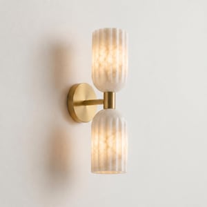 May include: A wall sconce featuring a circular brass mounting plate and a central arm that supports two vertically stacked, cylindrical, fluted glass shades with a translucent, marbled white finish that emits a soft, warm glow.