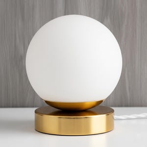 May include: A modern table lamp featuring a white globe shade atop a gold-colored base. The lamp has a round base and a gold-colored accent piece between the base and the shade. A white cord extends from the base.