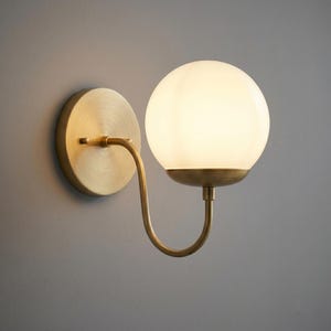 May include: A wall-mounted light fixture featuring a circular brushed brass backplate and a curved arm that supports a spherical, frosted white glass globe shade. The metal components have a warm, metallic finish, while the glass globe emits a soft, diffused light, creating a balanced and functional lighting piece for interior wall spaces.