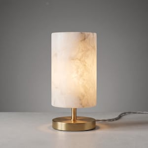 May include: A cylindrical table lamp featuring a translucent white and beige marbled stone shade. The lamp is supported by a circular brushed brass base and includes a matching fabric-covered power cord extending from the side of the base.