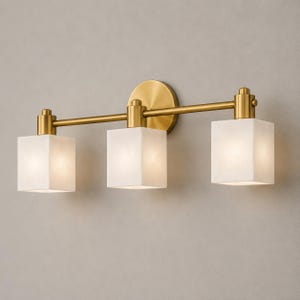 May include: A three-light wall fixture featuring a horizontal brass bar and a circular wall mount. Three square, white, frosted glass shades are attached to the bar, each emitting a soft, diffused light from within the cube-shaped glass housing.