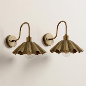 May include: Two antique brass wall sconces. Each sconce features a ruffled shade, a curved arm, and a round wall mount. The light bulbs are visible beneath the shades. The sconces are mounted on a neutral-colored wall.