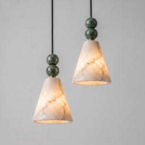 Op de afbeelding: Twee hanglampen met conische witte marmeren kappen met zichtbare aders. Elke lamp hangt aan een dunne zwarte stang en is voorzien van twee gestapelde, gepolijste groene marmeren bollen boven de conische kap.