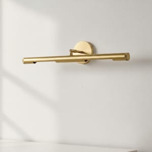 Peut inclure: Un luminaire doré pour tableau. Le luminaire est composé d'une base circulaire et d'une barre horizontale aux extrémités arrondies. La lumière est fixée sur un mur blanc. Le luminaire est en métal.