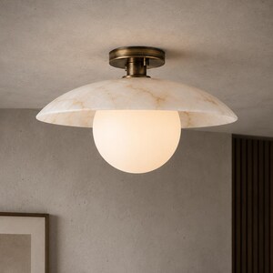 May include: A ceiling light fitting featuring a wide, shallow, marble-patterned dome shade in cream and tan tones, centred with a spherical frosted white glass globe. The fitting is mounted to the ceiling with a dark antique brass finish base.