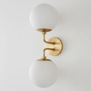 May include: A modern wall sconce featuring two spherical, frosted glass shades. The fixture has a brass finish with curved arms and a circular backplate. The design is minimalist and elegant, suitable for various interior styles.