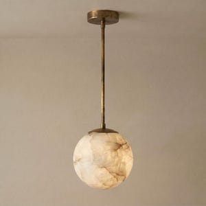 May include: A pendant light featuring a spherical shade made of translucent, marbled stone with cream and brown veining. The shade is suspended from a thin, vertical metal rod connected to a circular ceiling canopy, all finished in a muted antique brass.
