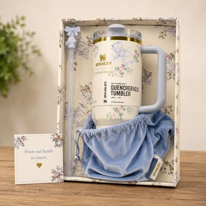 Pu&ograve; includere: Un bicchiere Stanley Quencher H2.0 in una confezione regalo. Il bicchiere color crema presenta un design floreale e un manico azzurro. La scatola include una cannuccia abbinata, un sacchetto di velluto blu e una carta con "Dalle nostre mani alle vostre".