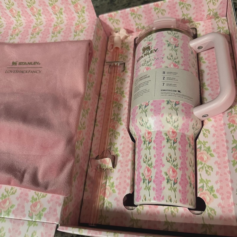 Pu&ograve; includere: Un bicchiere Stanley a fantasia floreale con manico, presentato in una scatola abbinata. Il bicchiere presenta un design a rose rosa e bianche. La scatola include una cannuccia rosa e un panno rosa. Il marchio "Stanley" &egrave; visibile.