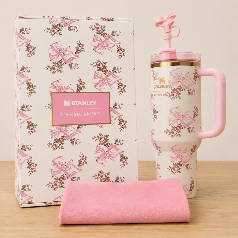 May include: A white Stanley tumbler with a pink bow and floral pattern, a matching pink handle, and a pink bow-shaped topper. The tumbler is next to a matching gift box and a pink cloth. The box has the text "Stanley Loveshackfancy".