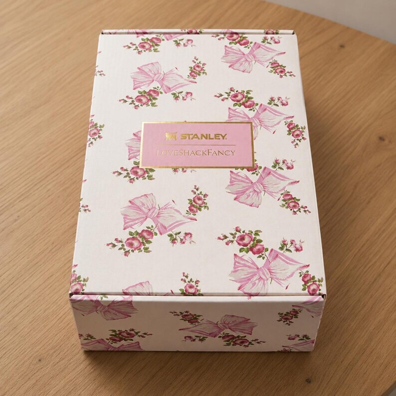 May include: A white box with a floral pattern of pink bows and roses. The box has a rectangular pink label with the text "STANLEY" and "LOVESHACKFANCY" in a gold frame. The box is on a light brown wooden surface.