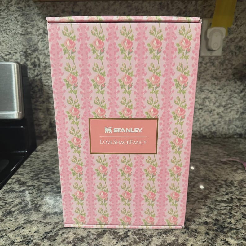 Pu&ograve; includere: Una scatola rosa con un motivo floreale di rose rosa e foglie verdi. La scatola ha un'etichetta rettangolare con il testo "STANLEY" e "LOVESHACKFANCY". La scatola &egrave; appoggiata su un piano di lavoro grigio.
