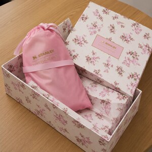 May include: A gift box with a floral pattern in pink and white, containing a pink drawstring bag. The bag has the text "STANLEY LOVESHACKFANCY" printed on it. The box is open, revealing the contents.