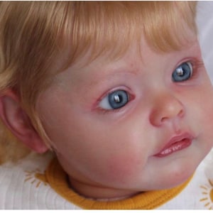 Puede incluir: Primer plano de una muñeca bebé con piel clara, ojos azules y cabello rubio. La muñeca lleva una camisa blanca con cuello amarillo y un diseño de sol. La cara de la muñeca tiene una apariencia realista con rasgos detallados.