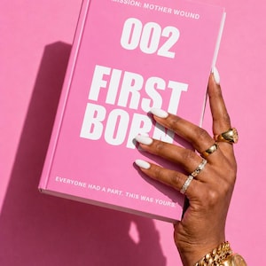 May include: A pink book titled "002 FIRST BORN" with the text "MISSION: MOTHER WOUND" and "EVERYONE HAD A PART. THIS WAS YOURS." in white. A hand with white nails and gold rings holds the book against a pink background. A gold watch and bracelet are also visible.