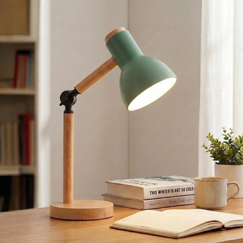 May include: A green desk lamp with a wooden arm and base. The lamp has a round, adjustable head and a warm light. The base is circular and made of wood. The lamp is on a wooden desk with books and a mug.