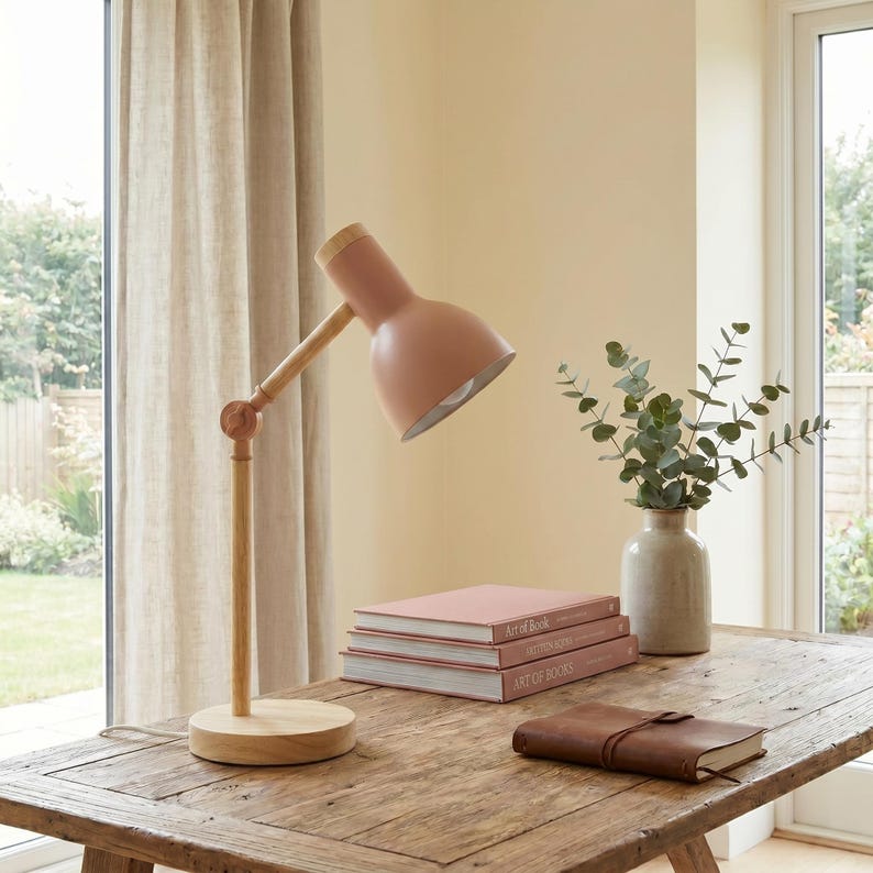 May include: A pink desk lamp with a wooden arm and base sits on a rustic wooden table. The lamp is angled towards a stack of pink books. A small vase with greenery and a leather-bound journal are also on the table.
