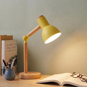 May include: A yellow desk lamp with a wooden base and arm. The lamp has a light bulb that is turned on. The lamp is on a wooden desk with a book, glasses, and a small blue ceramic pot with pens.
