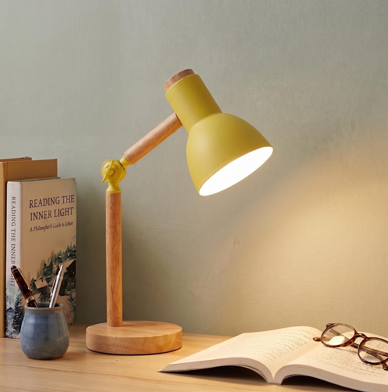 May include: A yellow desk lamp with a wooden base and arm. The lamp has a light bulb that is turned on. The lamp is on a wooden desk with a book, glasses, and a small blue ceramic pot with pens.