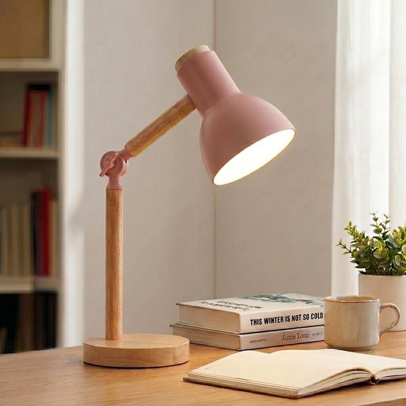 May include: A pink desk lamp with a wooden arm and base. The lamp's head is angled downward, illuminating the desk. The lamp is next to a stack of books and a mug. The lamp's design is modern and minimalist.