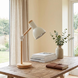 May include: A white desk lamp with a wooden arm and base sits on a wooden table. The lamp has a rounded shade and a light bulb. A stack of books and a leather-bound journal are also on the table. A vase with greenery is also on the table.