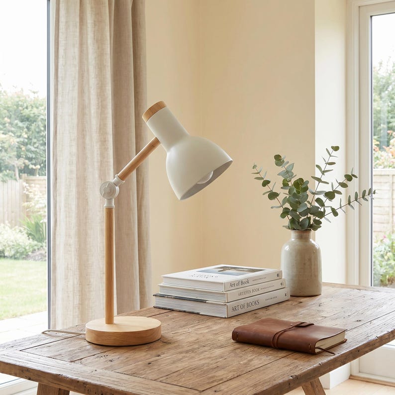 May include: A white desk lamp with a wooden arm and base sits on a wooden table. The lamp has a rounded shade and a light bulb. A stack of books and a leather-bound journal are also on the table. A vase with greenery is also on the table.