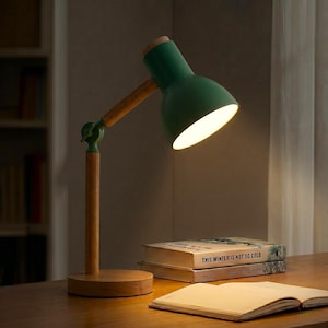 May include: A green desk lamp with a wooden arm and base is illuminated. The lamp is positioned over a wooden desk, with two books and an open notebook visible. The lamp's shade is pointed downward, casting a warm glow.