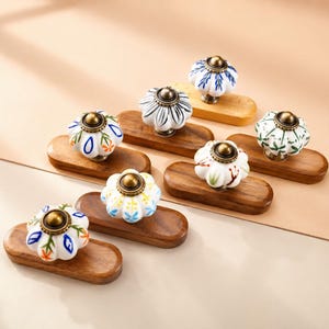 May include: A collection of decorative ceramic cabinet knobs with floral designs in various colors, including white, blue, green, and yellow. Each knob is mounted on a small, rectangular wooden base. The knobs have a brass-colored metal base.