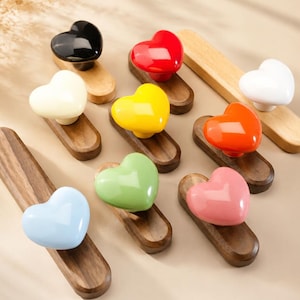 Puede incluir: Una colección de nueve pomos en forma de corazón en varios colores brillantes, incluyendo negro, rojo, blanco, amarillo, naranja, azul claro, verde claro y rosa. Cada corazón está montado sobre una base de madera alargada y lisa.