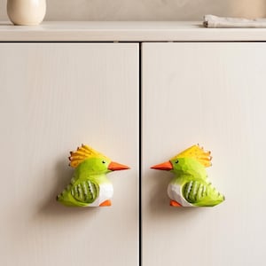 Könnte beinhalten: Zwei vogelförmige Schrankknöpfe an einem weißen Schrank. Jeder Knopf ist in leuchtendem Grün mit einem gelben Kamm, einem orangefarbenen Schnabel und weißen Details an den Flügeln bemalt. Die Vögel sind einander zugewandt und dienen als Griffe.