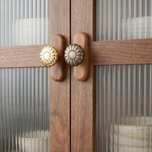 Puede incluir: Primer plano de un armario de madera con dos puertas. Cada puerta tiene un pomo decorativo en forma de flor. Las puertas del armario son de madera y tienen paneles de vidrio con detalles verticales y acanalados. Los pomos son dorados y bronce.