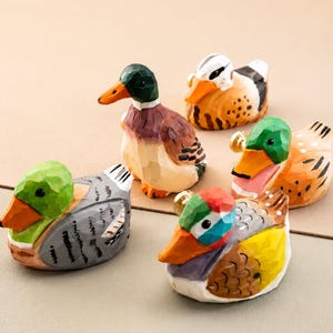 May include: A collection of five small, hand-carved wooden duck figurines. Each duck features distinct, vibrant painted details, including shades of green, orange, brown, grey, and yellow, with visible carved textures across their surfaces.