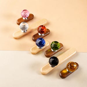 May include: A collection of seven wall hooks, each featuring a faceted, spherical crystal-like knob in various colors including pink, clear, orange, blue, green, black, and yellow, mounted on elongated, rounded wooden bases in light and dark tones.