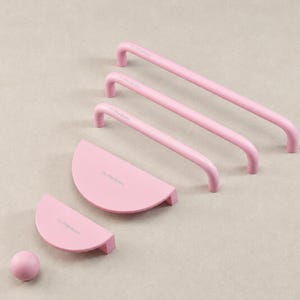 May include: A collection of matte pink hardware including three long tubular cabinet pulls, two semi-circular drawer pulls, and one small spherical knob. Each piece features the subtle embossed text "The Paper Room" on its surface.